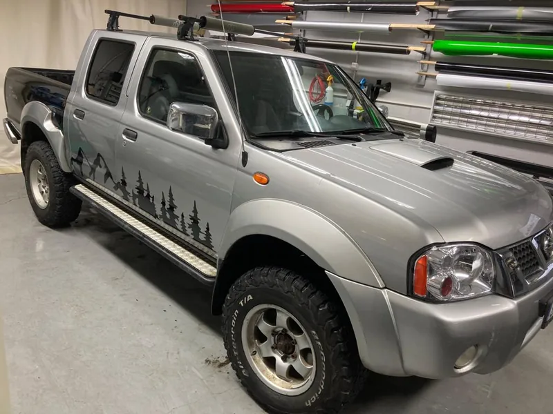 Stříbrný Nissan Navara s černou siluetou lesa podél boků