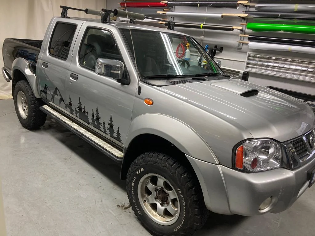Stříbrný Nissan Navara s černou siluetou lesa podél boků