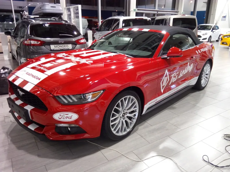 Červený Ford Mustang s bílým reklamním polepem Tři sestry zepředu a z boku